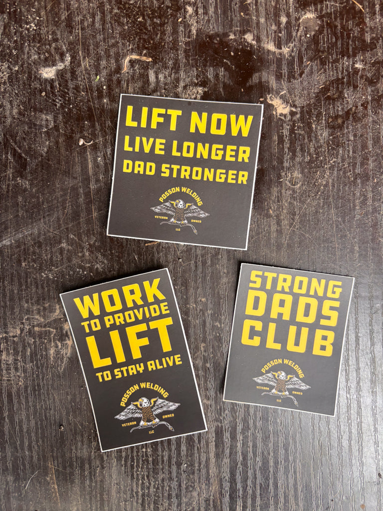 Three motivational stickers on a wooden surface with text like 'Lift Now Live Longer Dad Stronger' and 'Strong Dads Club'.