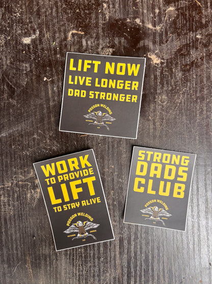Three motivational stickers on a wooden surface with text like 'Lift Now Live Longer Dad Stronger' and 'Strong Dads Club'.